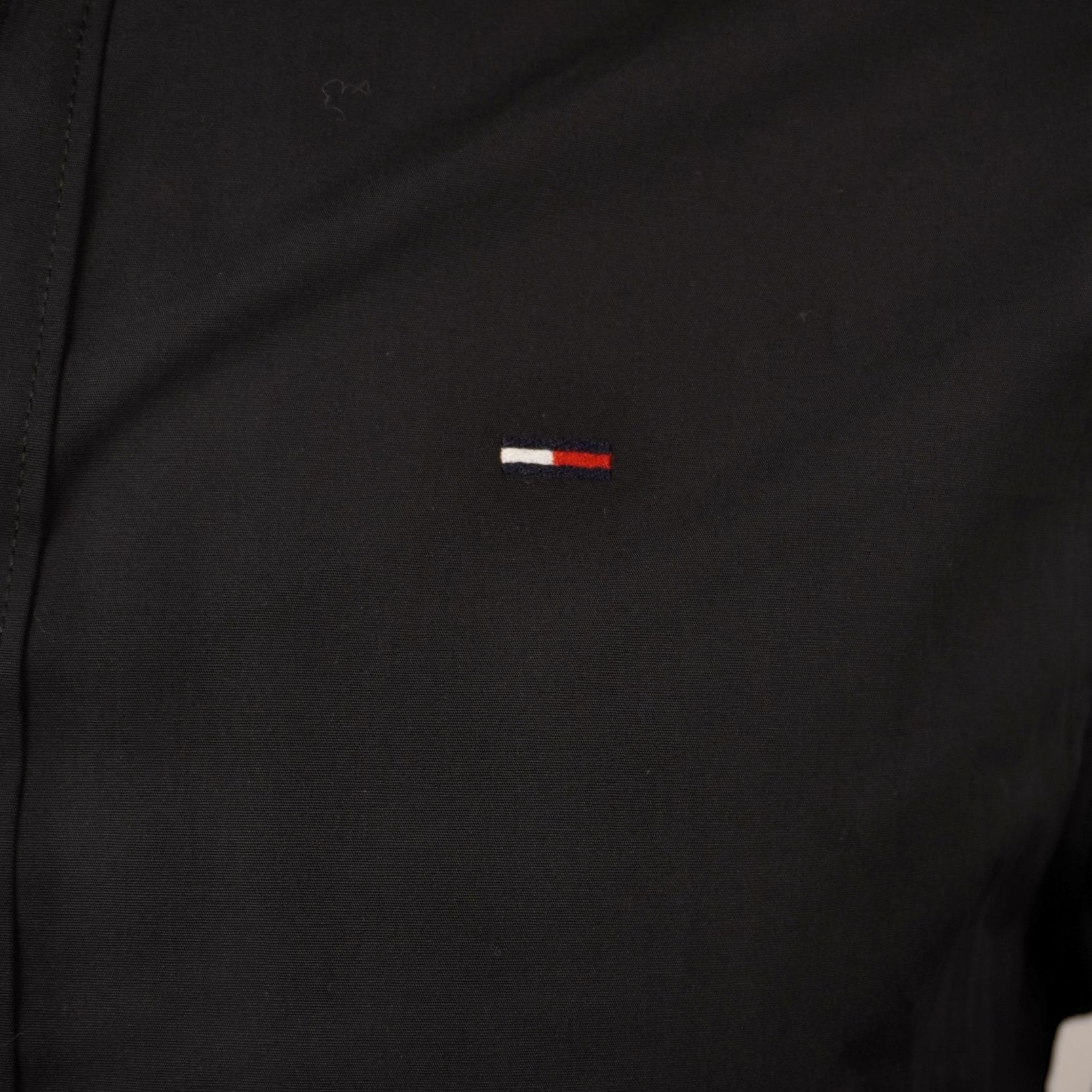 Inquadratura macro dell'iconico logo Flag di Tommy Jeans ricamato nei colori bianco, rosso e blu sul petto della camicia nera.