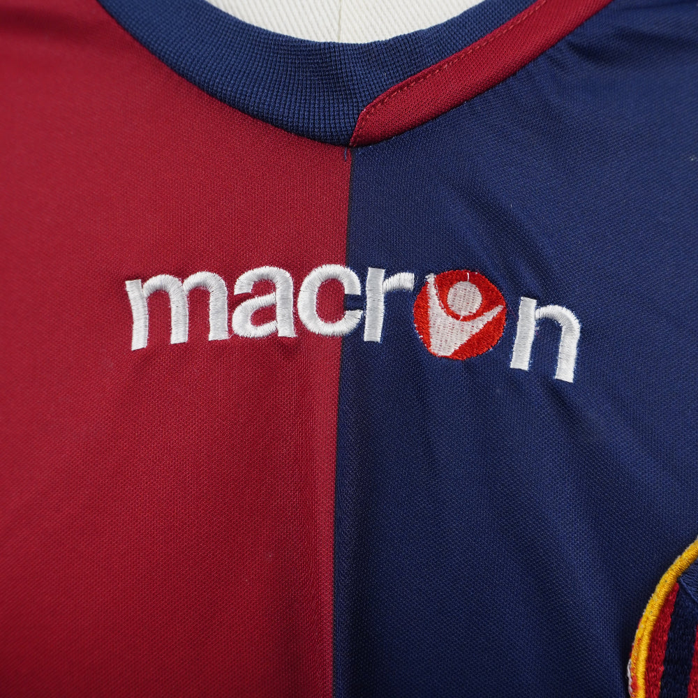Primo piano del logo Macron ricamato in bianco al centro del petto sulla maglia da calcio rossoblù