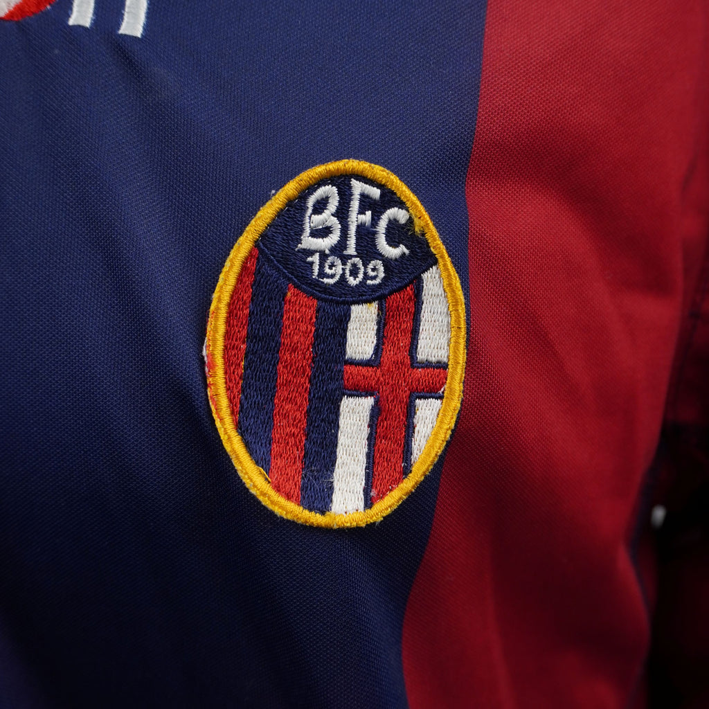 Inquadratura ravvicinata dello stemma ufficiale del Bologna FC 1909 ricamato sul petto della divisa casalinga