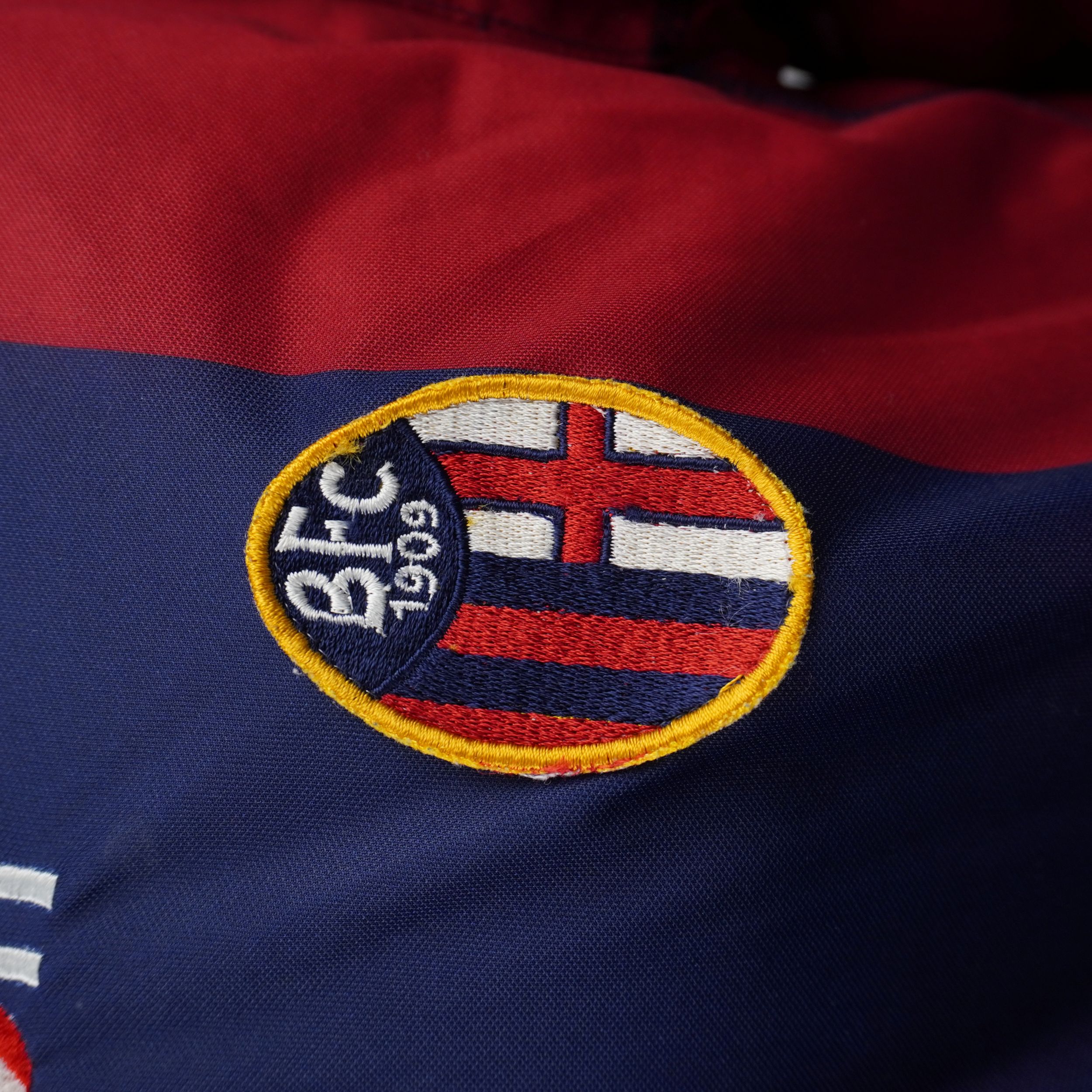 Inquadratura ravvicinata dello stemma ufficiale del Bologna FC 1909 ricamato sul petto della divisa casalinga