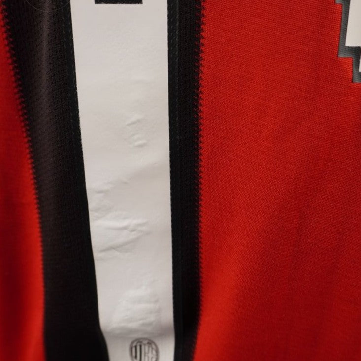 Dettaglio ravvicinato della base del numero stampato sulla maglia del Milan, che mostra il piccolo logo societario rossonero inserito all'interno della cifra bianca.