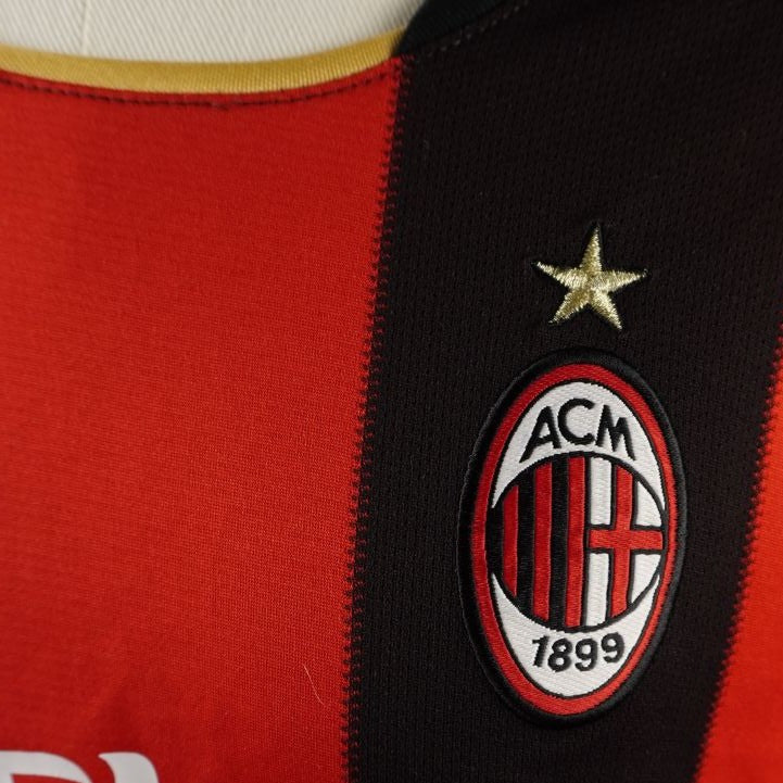 Primo piano dello stemma societario dell'AC Milan con la stella d'oro ricamato sul petto, lato cuore, su tessuto tecnico a righe rossonere.