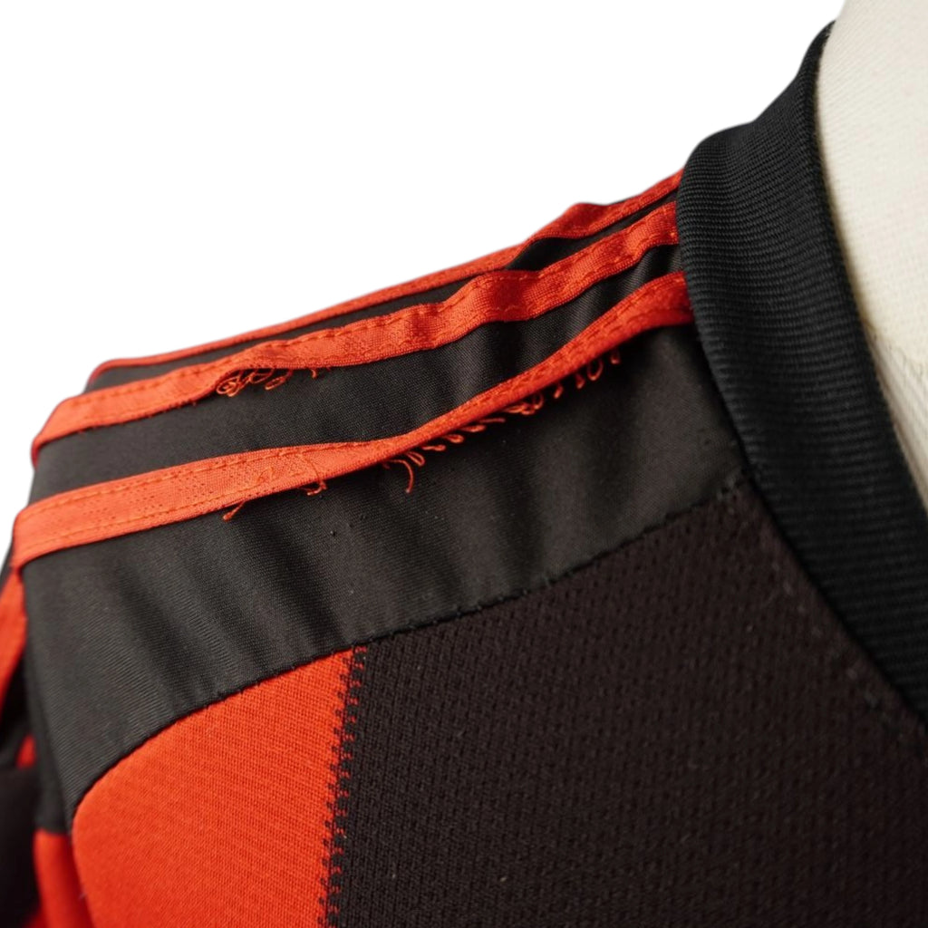 Primo piano ravvicinato della spalla della maglia del Milan che evidenzia sfilacciature e scuciture lungo i bordi delle tre strisce rosse Adidas applicate.