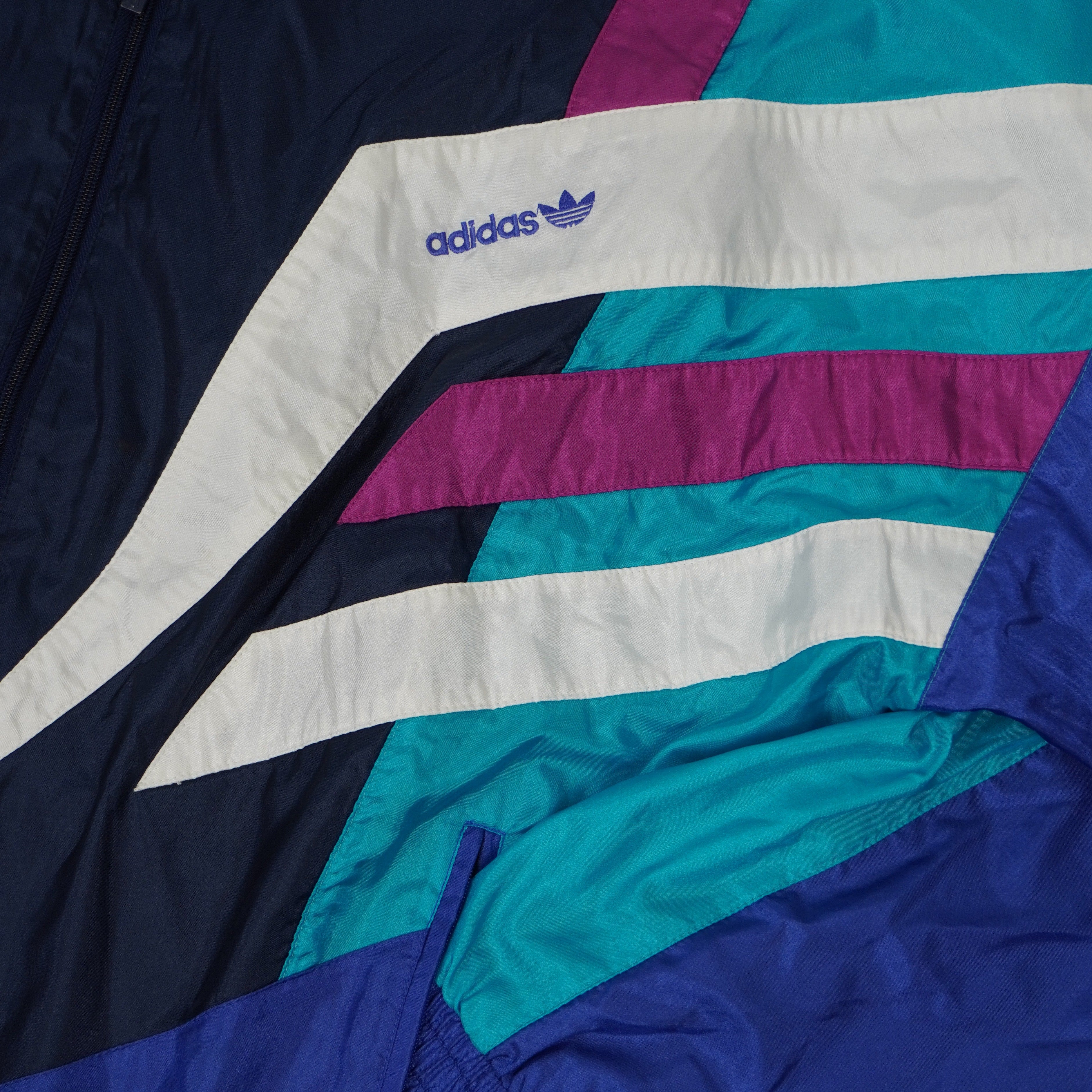 Dettaglio del ricamo logo Adidas Trefoil viola su pannello geometrico bianco anni novanta