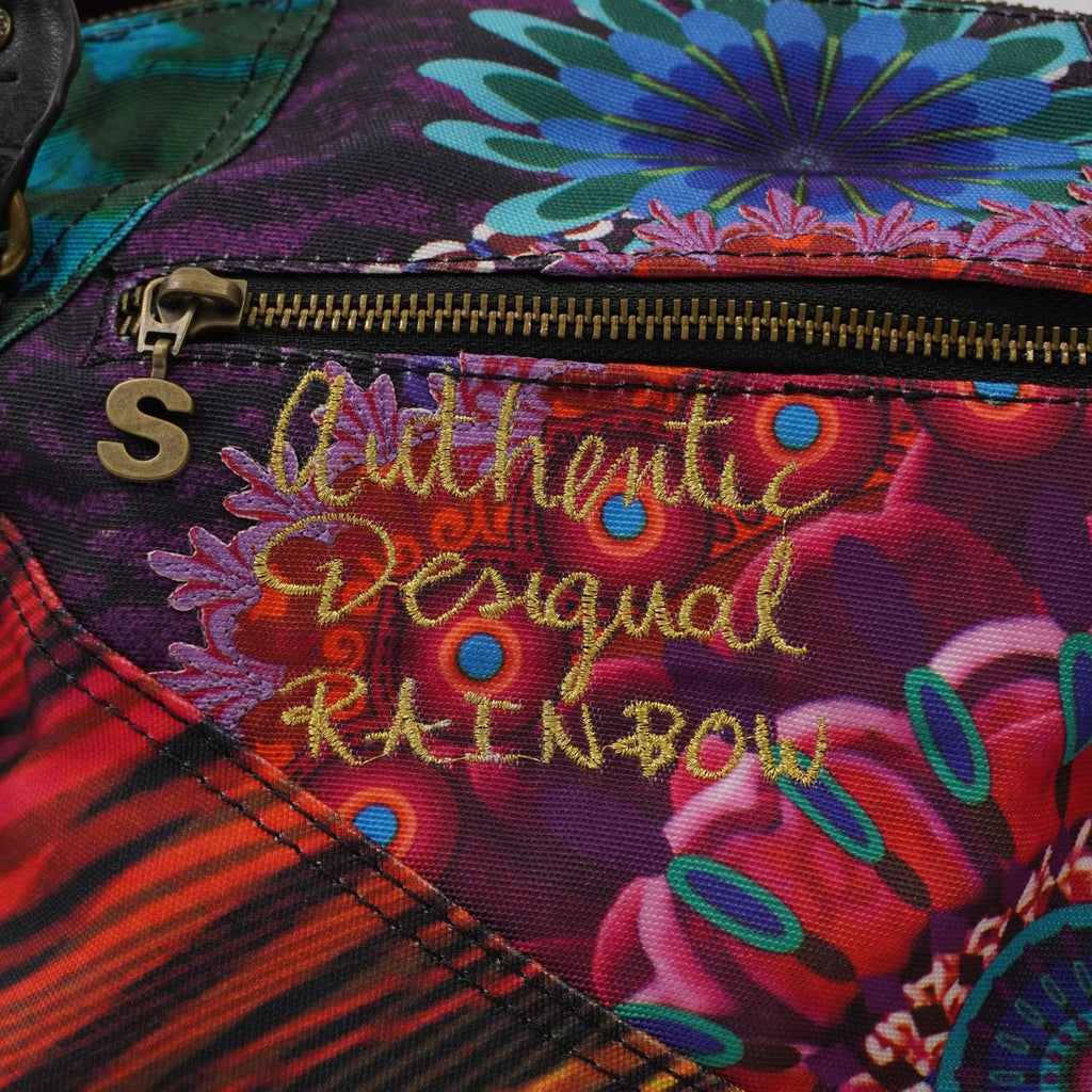 Primo piano del ricamo posteriore sulla borsa con la scritta "It's authentic Desigual brand" in filo dorato su tessuto a motivi floreali viola e blu