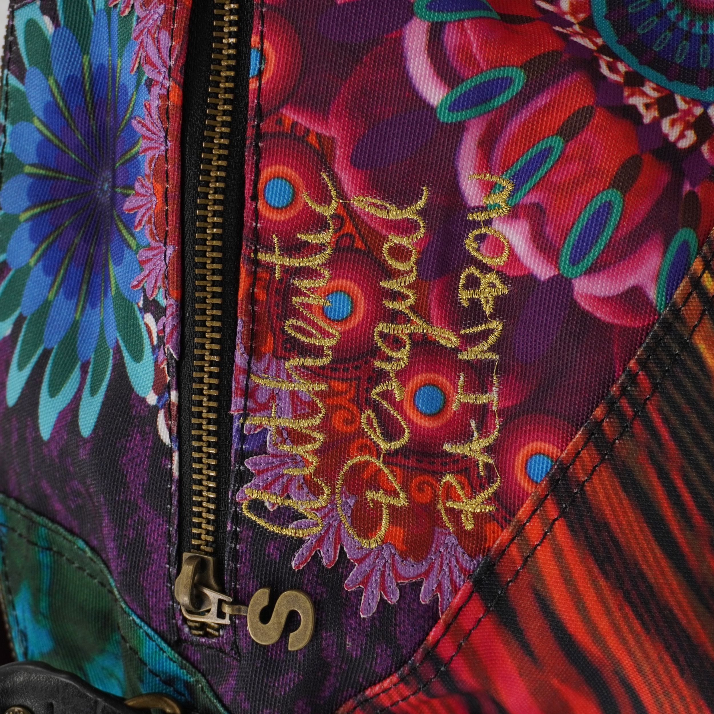 Primo piano del ricamo posteriore sulla borsa con la scritta "It's authentic Desigual brand" in filo dorato su tessuto a motivi floreali viola e blu