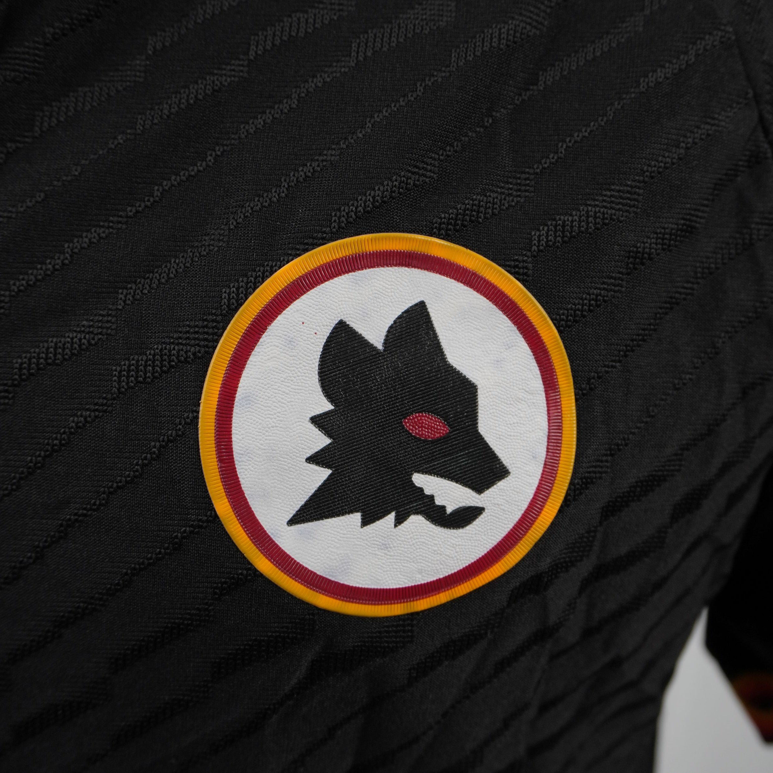 Inquadratura ravvicinata dello stemma gommato in rilievo del Lupetto di Piero Gratton sul petto della maglia nera della Roma